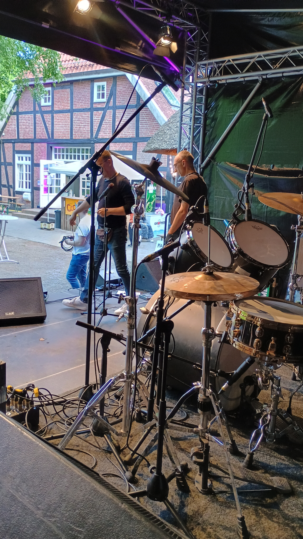 Ein Foto der Bühne beim Aufbau. Im Vordegrund sieht man das Schlagzeug, im Hintergrund die Vorderseite der Bühne, wo gerade zwei Männer Gitarren verkabeln. Ganz hinten ist ein Fachwerkhaus zu sehen.