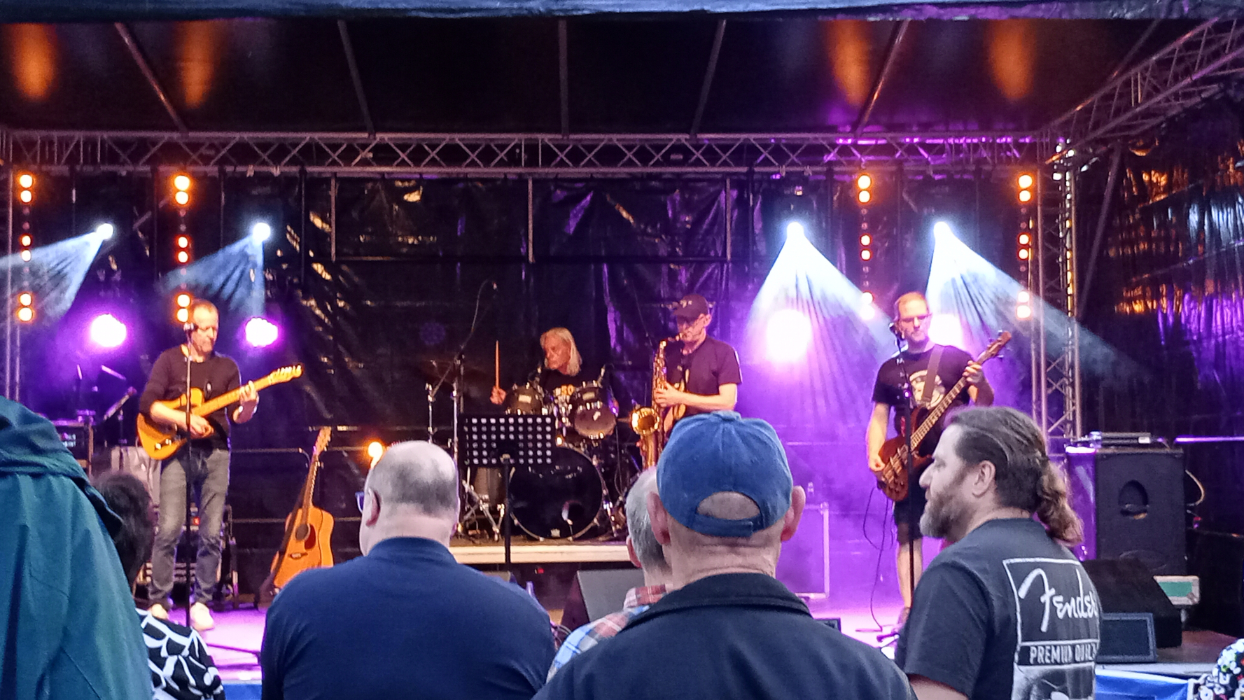 Ein Foto der Bühne mit der Band 'Ohrenblick'. Olli an der Gitarre, Andy am Schlagzeug, Carsten am Saxophon und Carsten am Bass.
