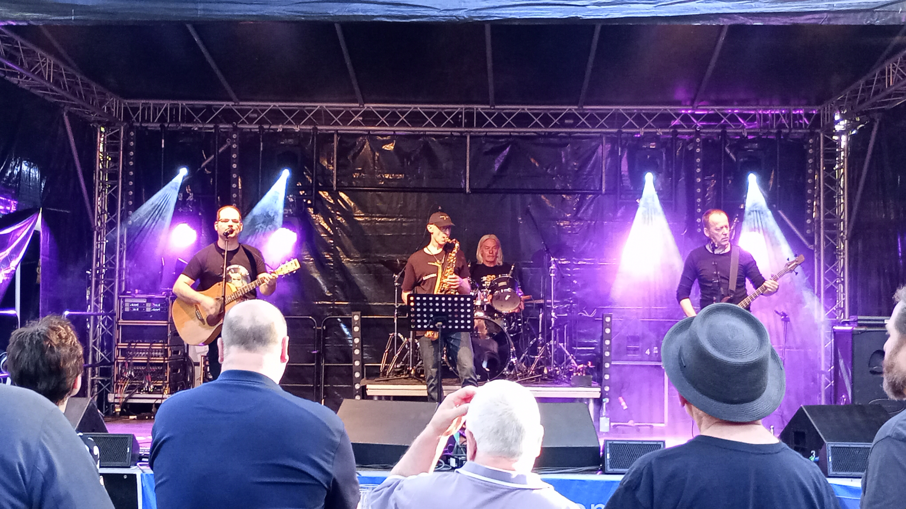 Ein Foto der Bühne mit der Band 'Ohrenblick'. Carsten an der Gitarre, Andy am Schlagzeug, Carsten am Saxophon und Olli am Bass.