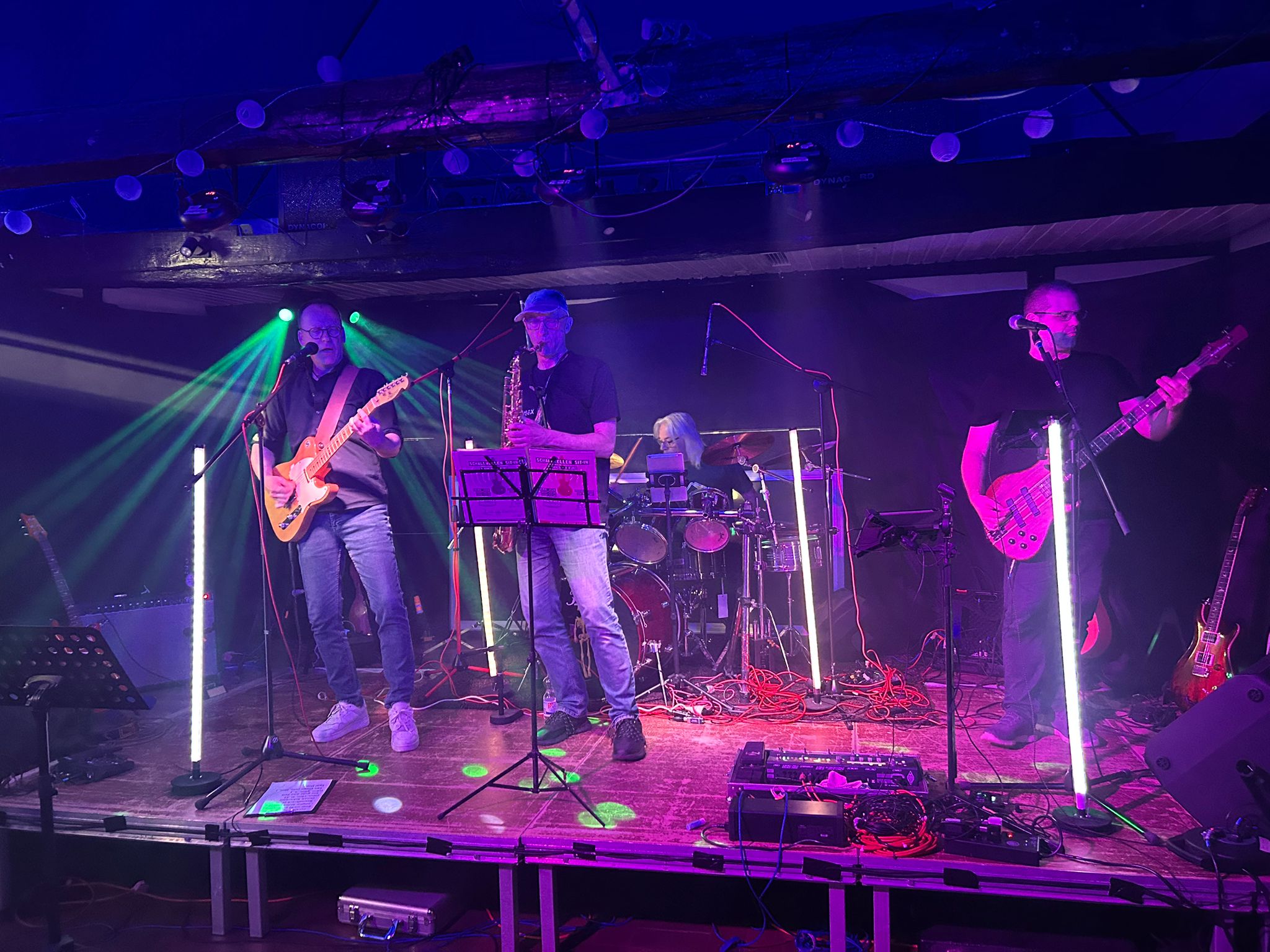 Ein Foto der Bühne mit der Band 'Ohrenblick'. Olli an der Gitarre, Andy am Schlagzeug, Carsten am Saxophon und Carsten am Bass.