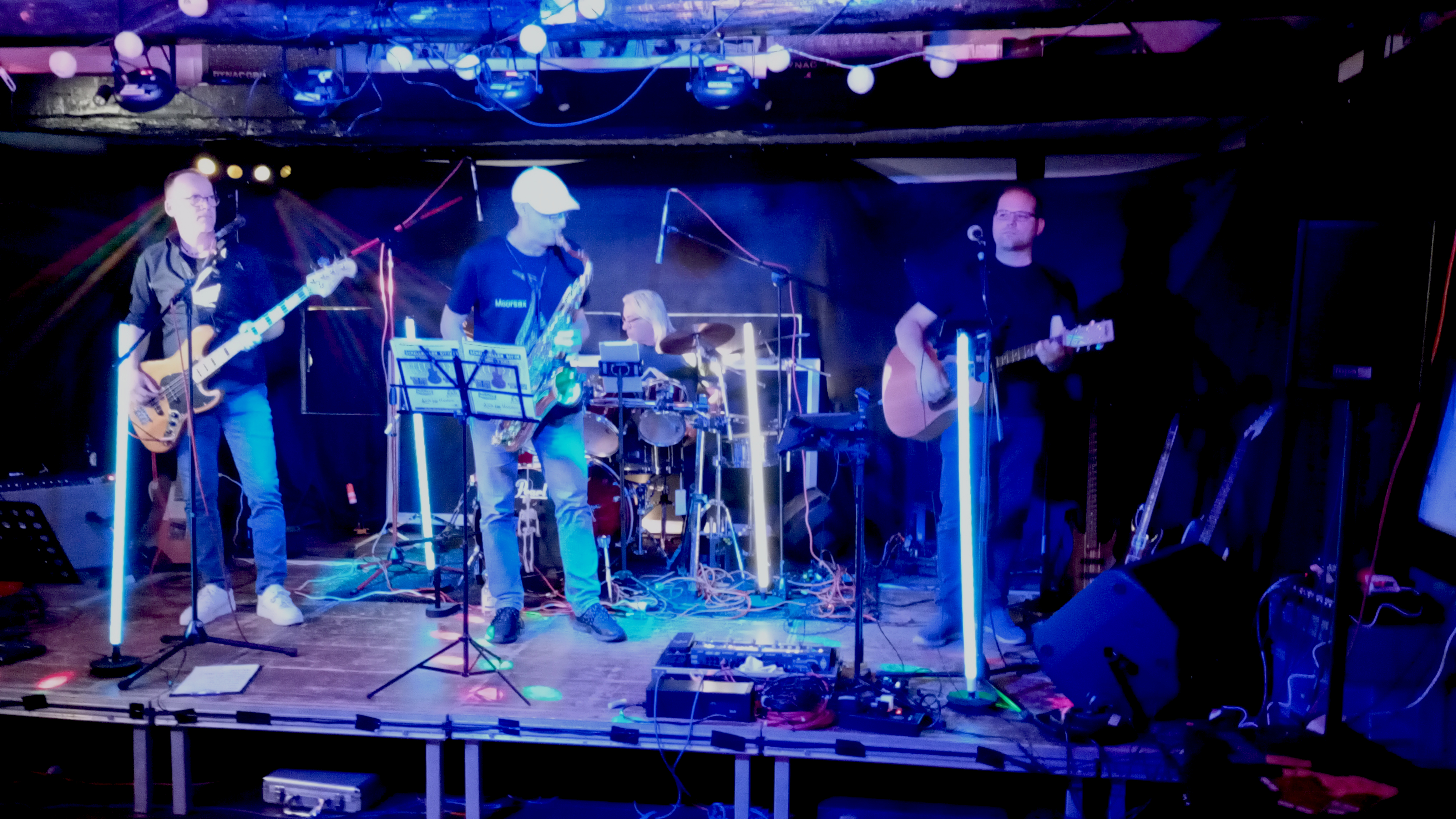 Ein Foto der Bühne mit der Band 'Ohrenblick'. Olli am Bass, Andy am Schlagzeug, Carsten am Saxophon und Carsten an der Gitarre.
