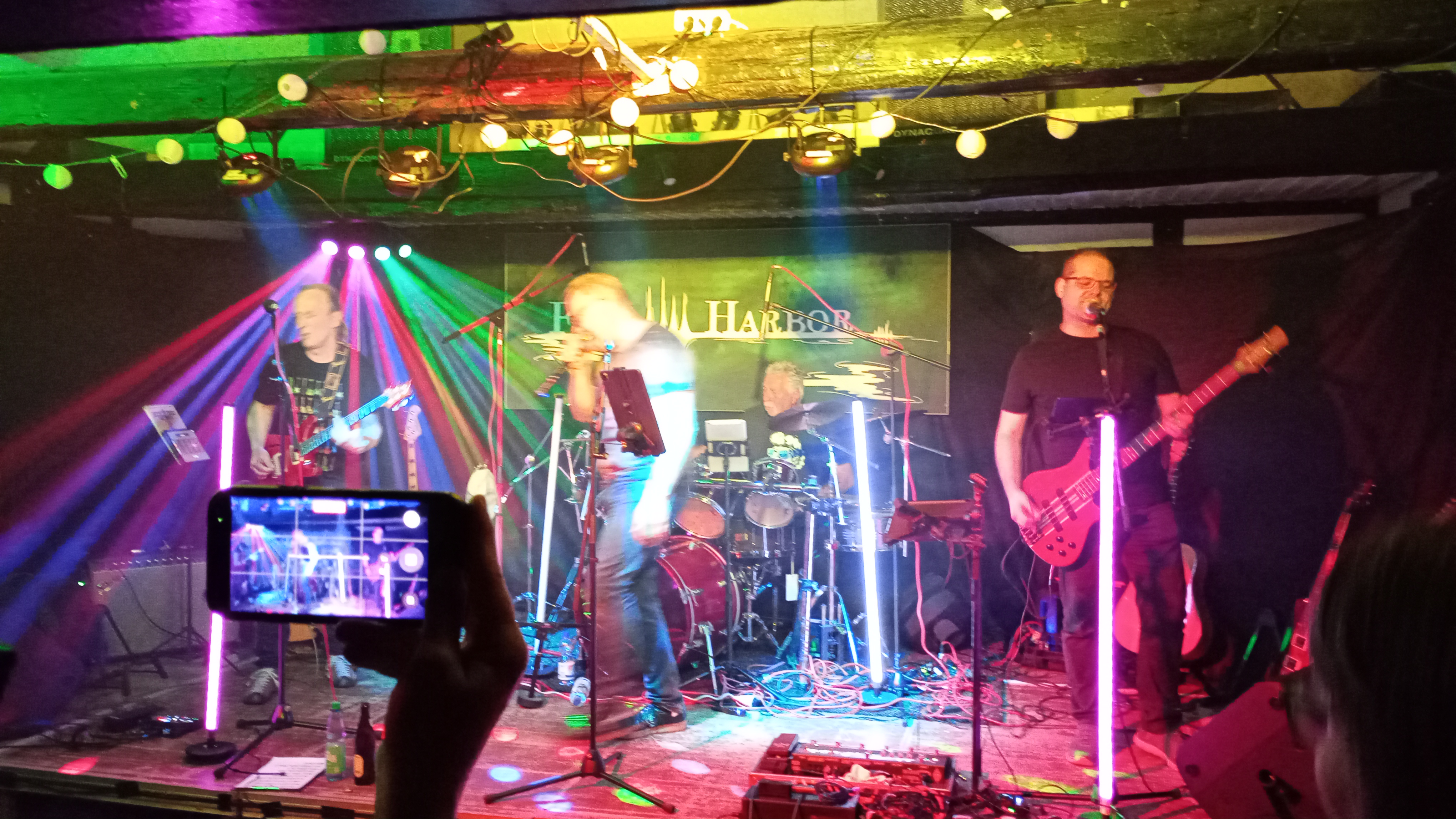 Ein Foto der Bühne mit der Band 'Fen Harbor'. Im Vordergrund sieht man, wie jemand mit einem Smartphone filmt.