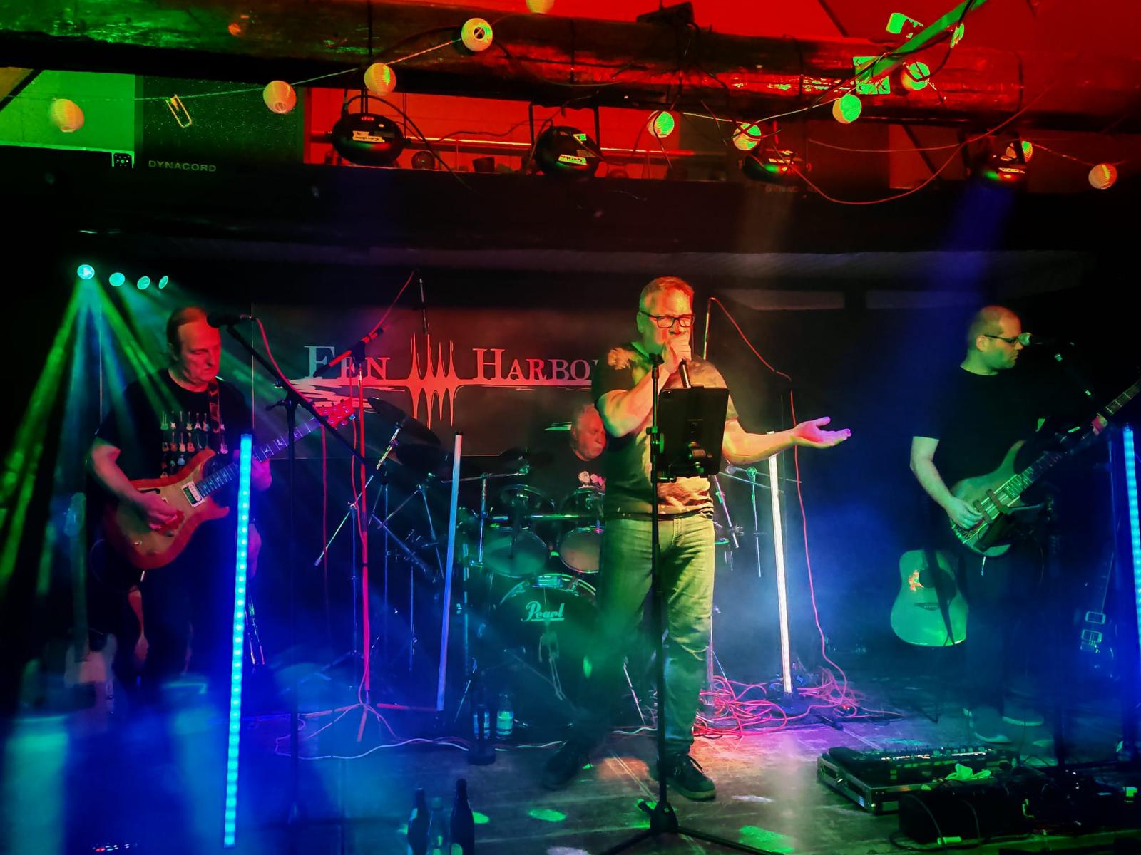 Ein Foto der Bühne mit der Band 'Fen Harbor'. Andreas an der Gitarre, Rüdiger am Schlagzeug, Vokler am Mikrofon und Carsten am Bass.